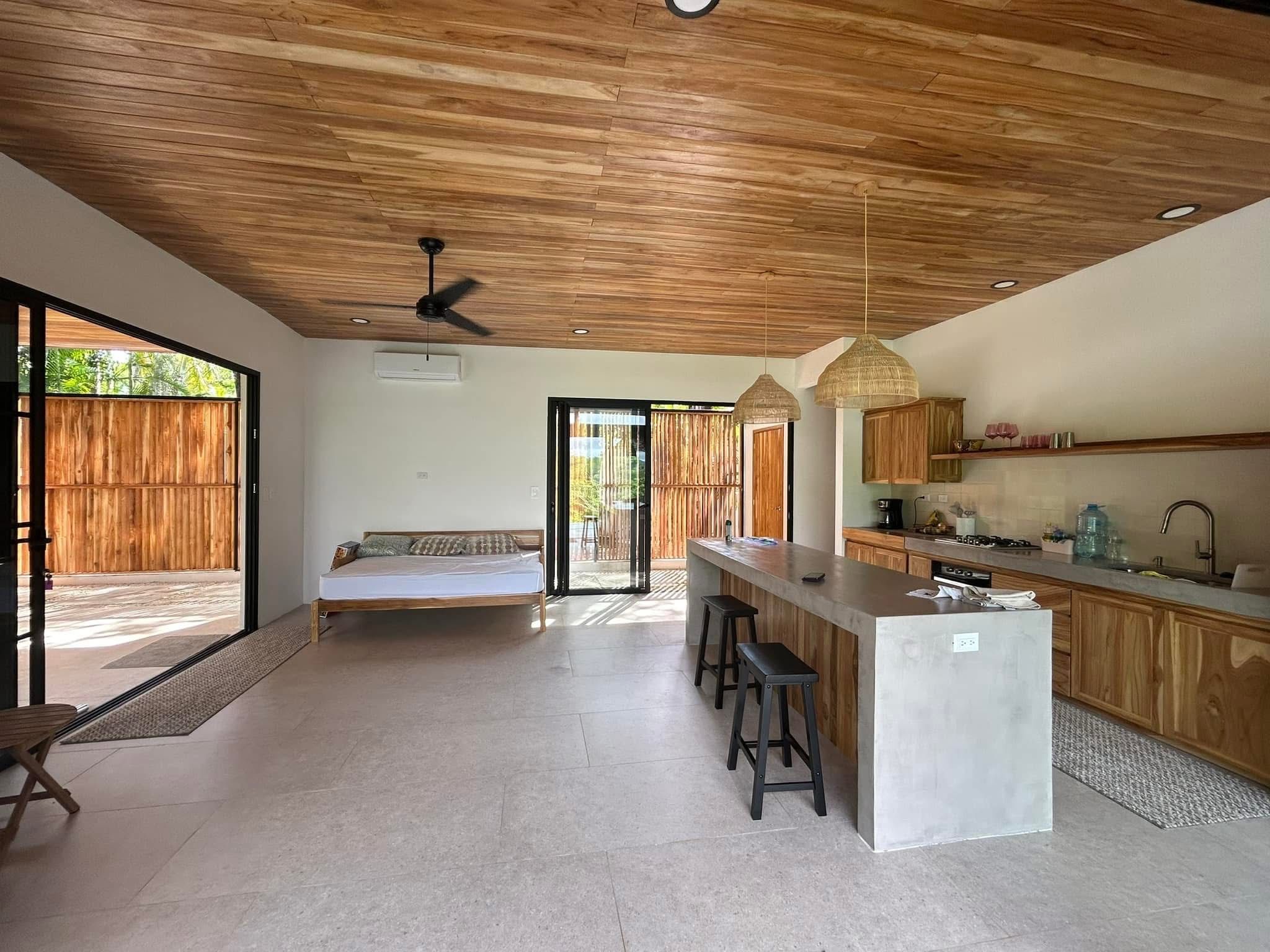 Open kitchen and dining in Casa Delicias with wood-slat ceiling, monolithic concrete island, and integrated bar seating.